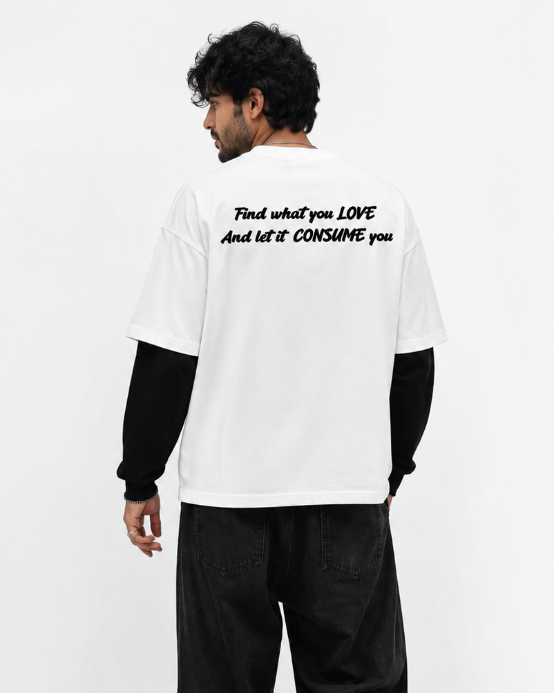 A man shown from the back wearing a white graphic t-shirt with black text, layered over a black long-sleeve shirt, black jeans, and white sneakers, against a plain white studio background.